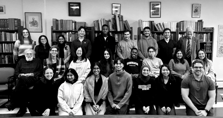 24 people, young adults and older, are grouped in front of a few full bookshelves. The photo is in black and white